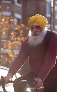 An older man wearing a yellow turban cycles down a sunny street