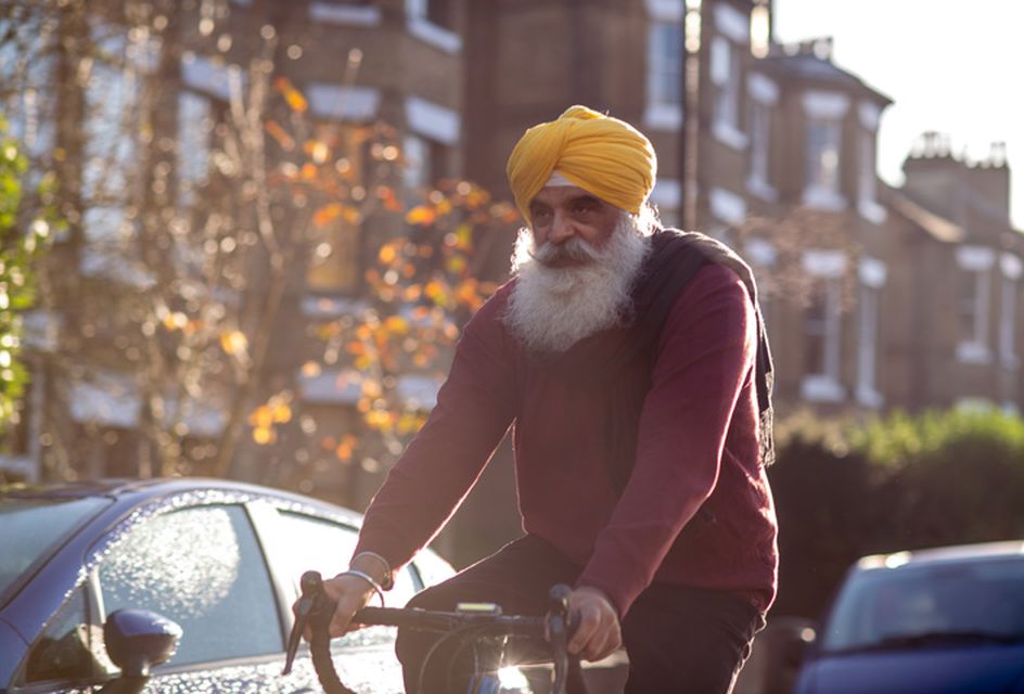 An older man wearing a yellow turban cycles down a sunny street