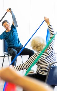 A man leads an exercise class for elderly people. He demonstrates stretching with an exercise band and the class copies.