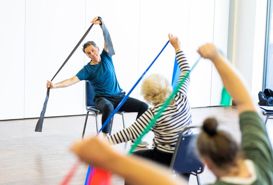 A man leads an exercise class for elderly people. He demonstrates stretching with an exercise band and the class copies.