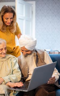 An older Asian couple are sat on a sofa looking at a laptop. A younger woman stands behind them, also looking at the laptop.