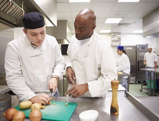 Two chefs are in an industrial kitchen chopping vegetables on a green chopping board.