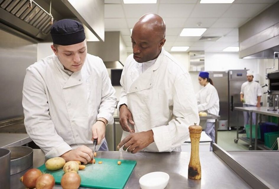 Two chefs are in an industrial kitchen chopping vegetables on a green chopping board.