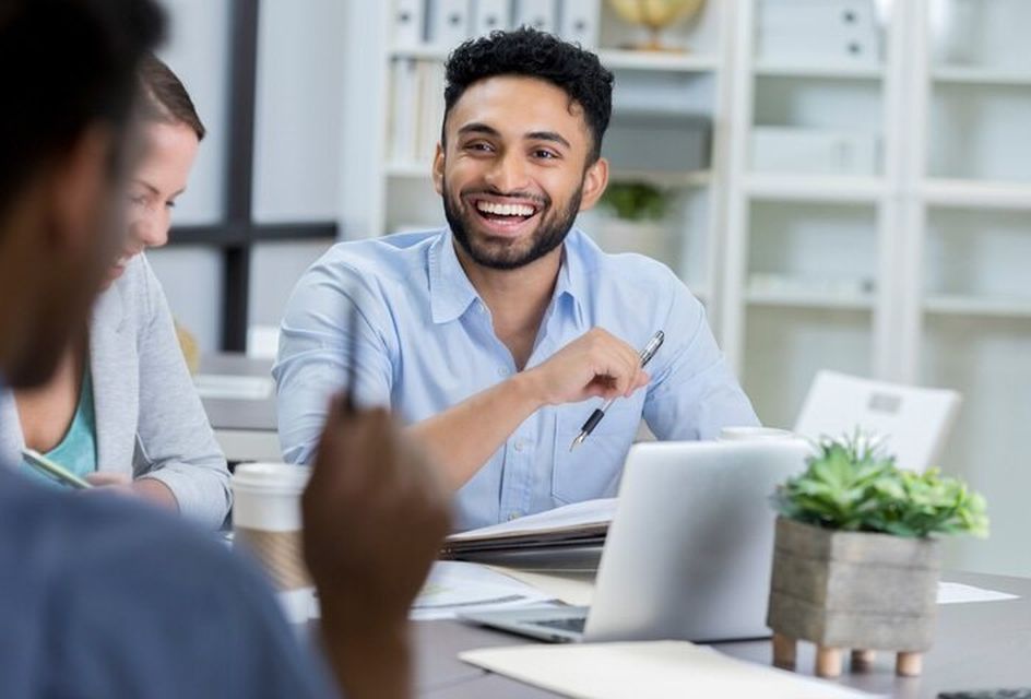 A young man smiles with colleagues while working on a laptop and notebook.