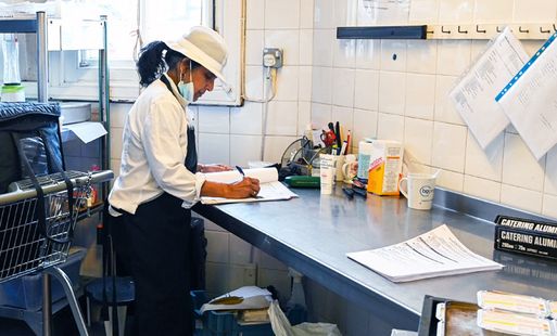 Catering staff member in uniform filling out paperwork at a stainless steel workstation in a commercial kitchen.