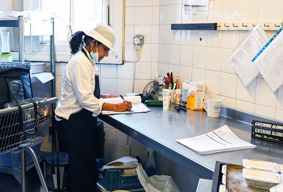Catering staff member in uniform filling out paperwork at a stainless steel workstation in a commercial kitchen.