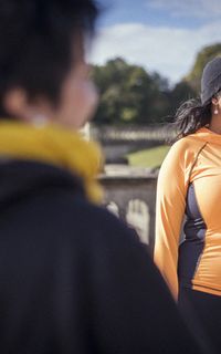 A black woman with long black hair, a black hair and an orange top stands in a park.