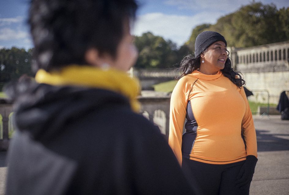 A black woman with long black hair, a black hair and an orange top stands in a park.
