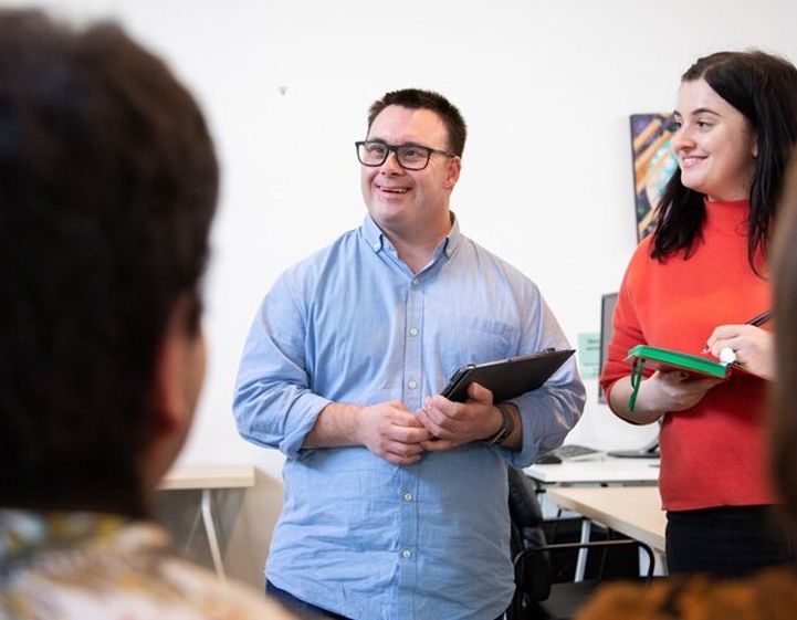 A man holding a clipboard smiles at his colleagues as they talk in an office setting. The man is white and wears blue shirt and glasses. Next to him is a woman with brown hair and a red top.
