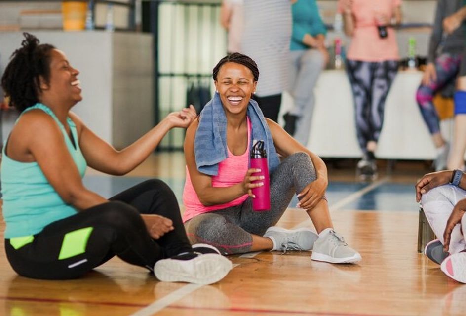 Two black women sit on the floor of a gym laughing and talking. They are wearing sports clothes and one has a towel around her neck.