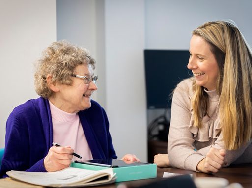 An elderly white woman with glasses writes on a notepad as she talks to a care worker