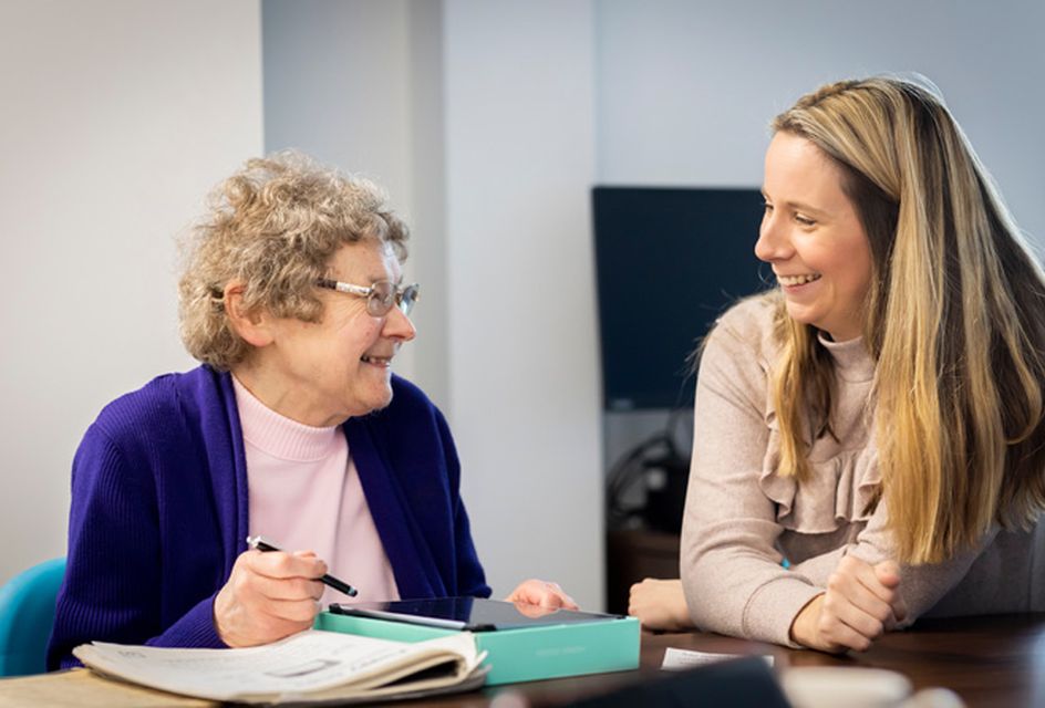 An elderly white woman with glasses writes on a notepad as she talks to a care worker