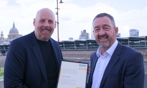 William Roberts and Chris Borardman CBE smiling while holding an RSPH award certificate on Blackfriars bridge, with London landmarks visible in the background