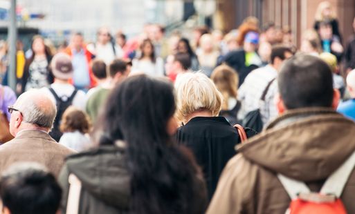 Large crowd of people walking in a busy urban area, viewed from behind.