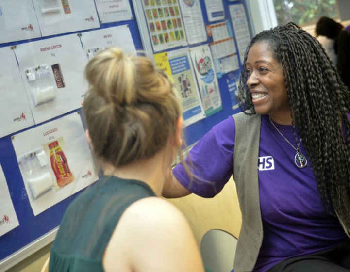 A woman wearing a purple NHS branded tshirt points to a wall of information on sugar while talking to another woman.