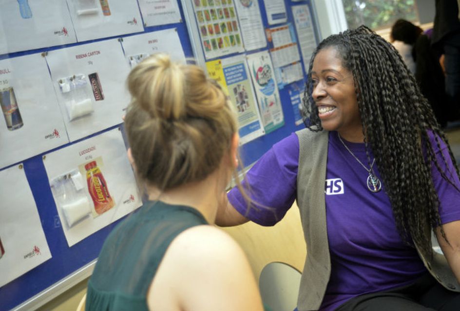 A woman wearing a purple NHS branded tshirt points to a wall of information on sugar while talking to another woman.