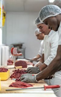 A line of chefs wearing aprons and hair nets chopping meat in a kitchen.