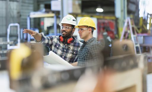 Two man wearing hard hats and safety goggles in a warehouse setting. One of the men is pointing to something out of shot.
