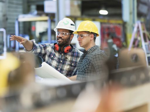 Two man wearing hard hats and safety goggles in a warehouse setting. One of the men is pointing to something out of shot.