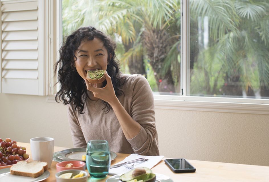 An Asian woman with black curly hair smiles while eating a piece of toast with avocado on it.
