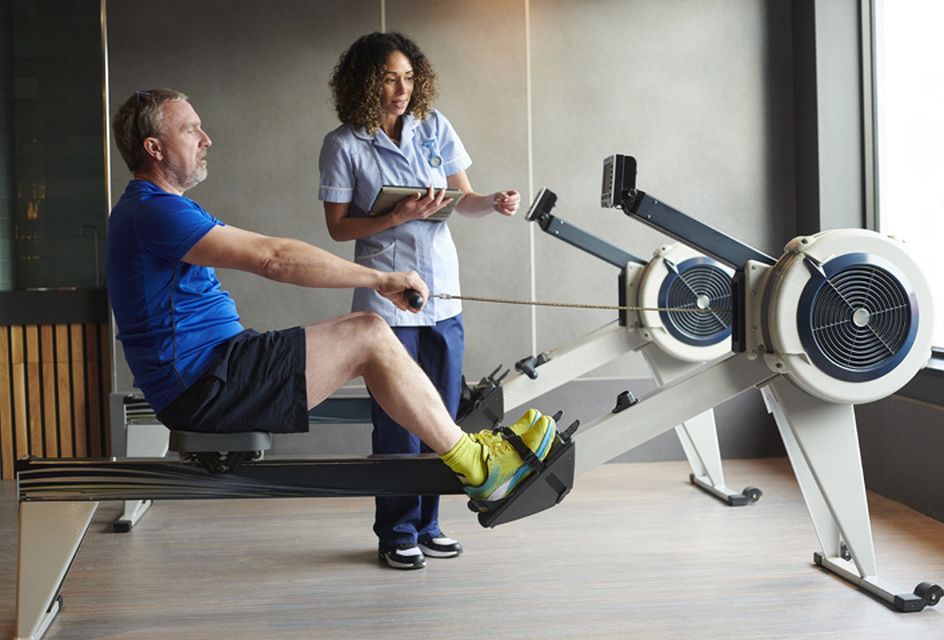 A physio assists a man as he uses a rowing machine.