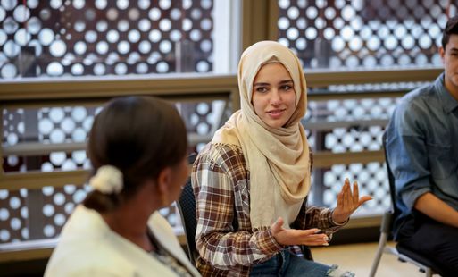 A young woman in engages in a group discussion, gesturing expressively. She sits alongside others in a modern, light-filled room.