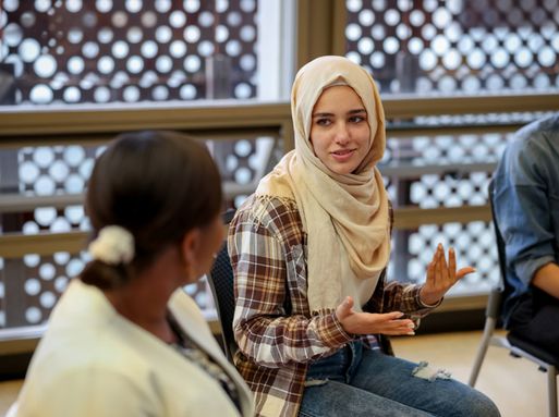 A woman wearing a hijab and plaid shirt speaks to a group of people.