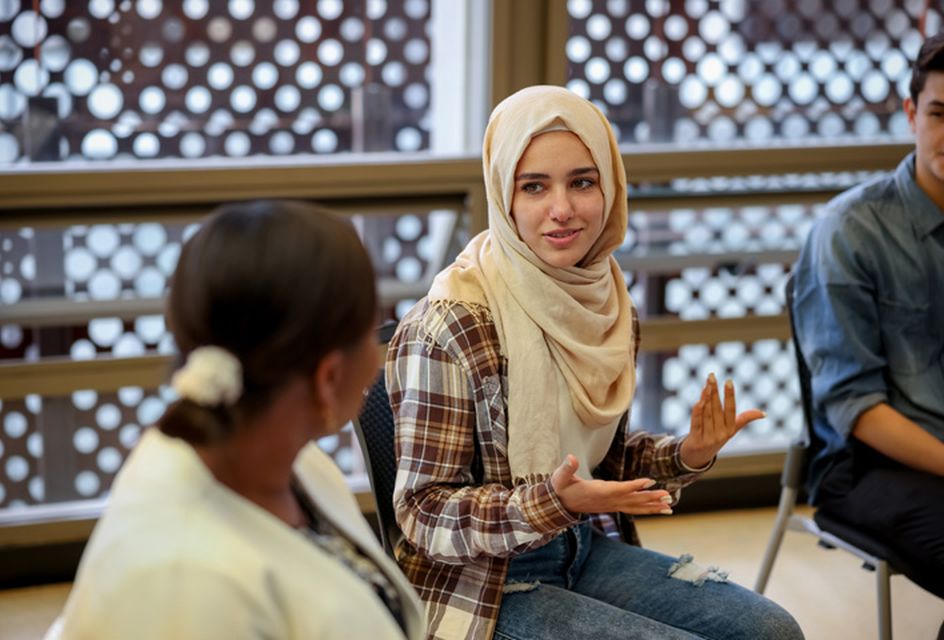 A young woman in engages in a group discussion, gesturing expressively. She sits alongside others in a modern, light-filled room.