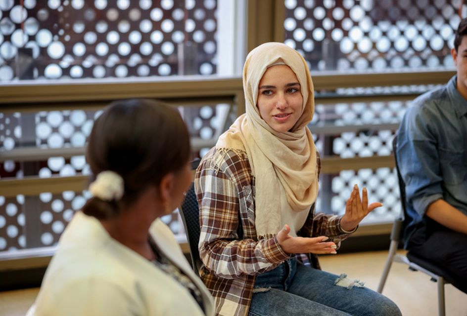 A woman wearing a hijab and plaid shirt speaks to a group of people.