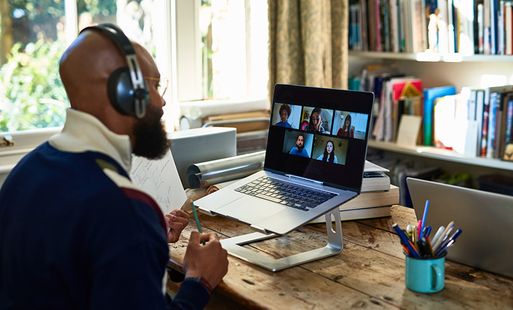 A man wearing headphones joins an online meeting on his laptop. On the laptop screen there are lots of faces in boxes.