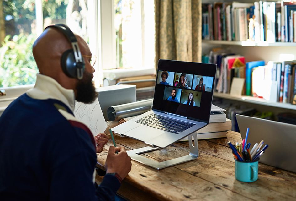 A man wearing headphones joins an online meeting on his laptop. On the laptop screen there are lots of faces in boxes.