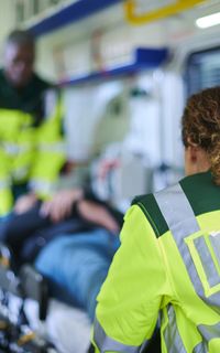 A paramedic wearing a high-vis that reads "ambulance" pushes a patient in a stretcher into an ambulance.