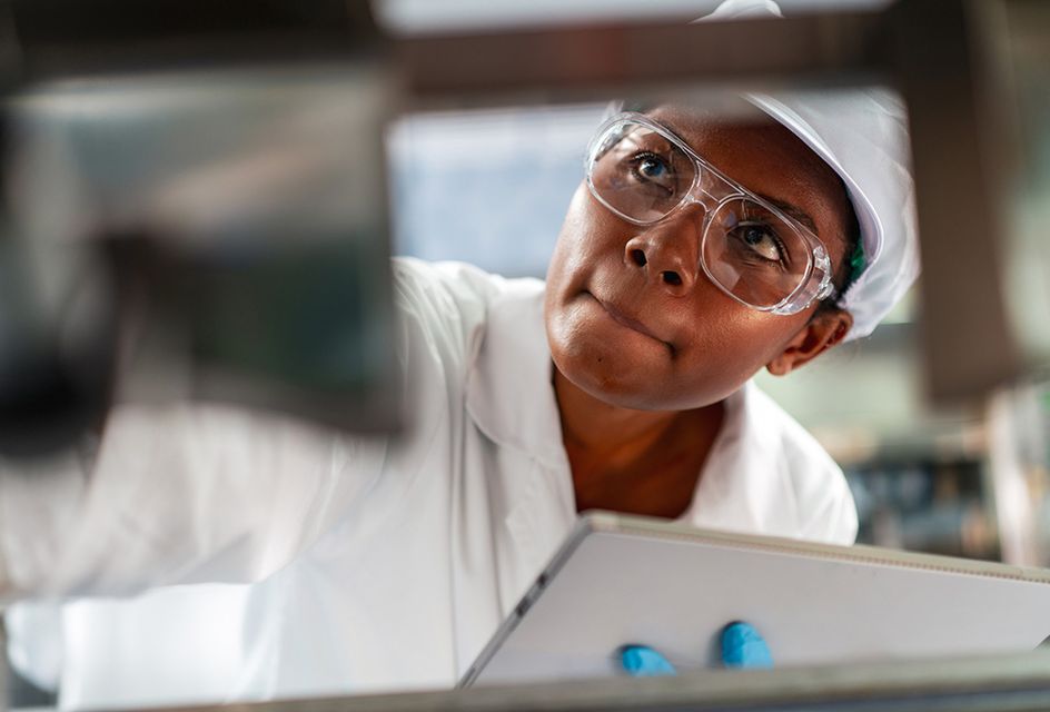 A woman in a white coat, hair net and goggles inspects a piece of machinery in a factory and takes notes on a clipboard.