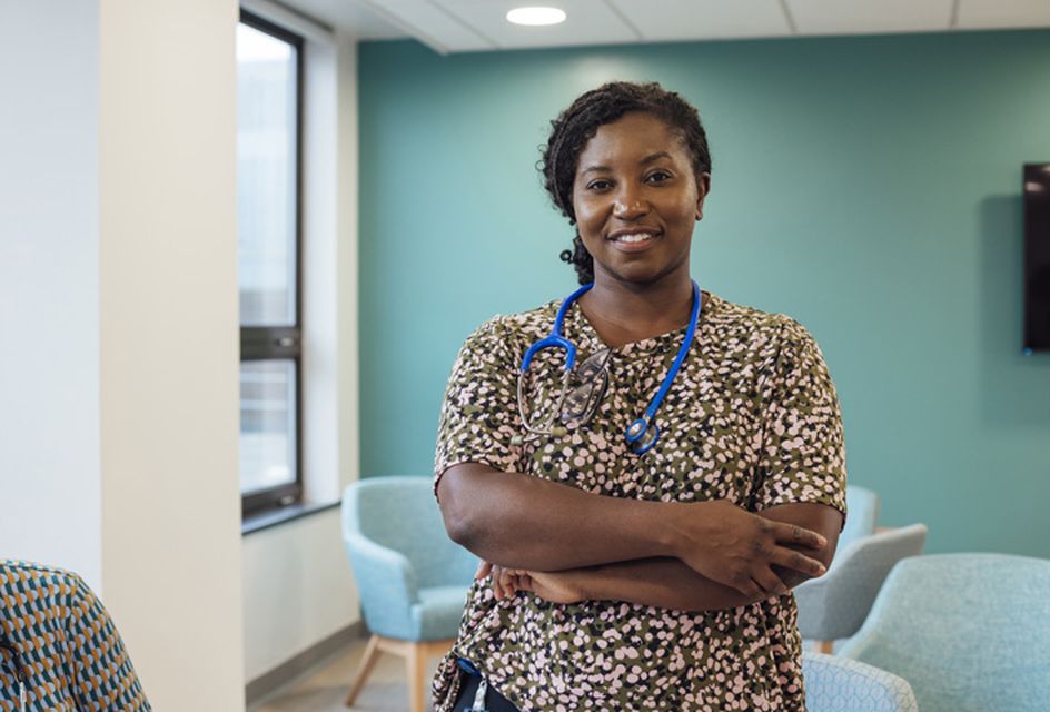 Female doctor wearing stethoscope with crossed arms smiling at the camera in a clinical waiting room.