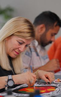 A white woman with blonde hair paints a colourful picture. Two others are in the background painting too.