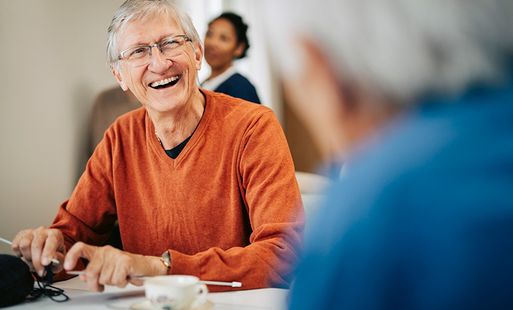 An elderly white man with grey hair, glasses and an orange jumper is smiling while knitting in a community setting.