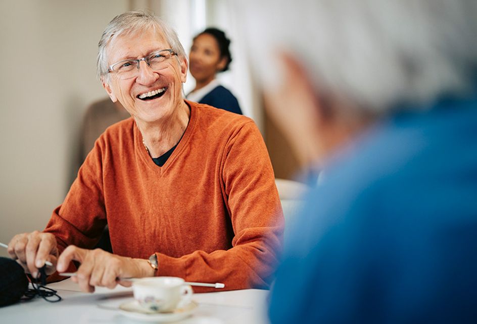 An elderly white man with grey hair, glasses and an orange jumper is smiling while knitting in a community setting.
