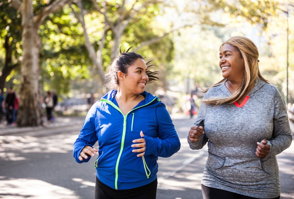 Two women jogging side by side smiling at each other on a sunny day with trees lining the street.
