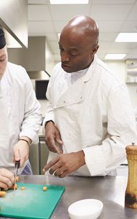 A chef in a white coat supervises another chef as he chops onions.