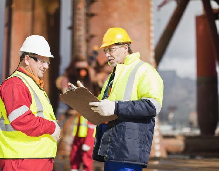 Two white men wearing high vis jackets and hard hats talk on a construction site.