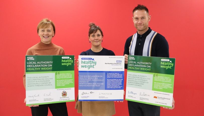 Three people stand smiling against a red background, each holding a "Healthy Weight Declaration" sign. The tone is positive and collaborative.