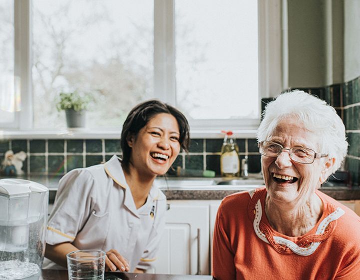 An elderly white woman with grey hair and glasses laughs with her carer, and younger woman with brown hair.