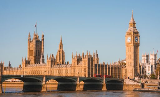 The Houses of Parliament and Westminster Bridge in full view in sunlight with two London buses going over the bridge.