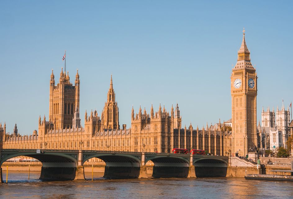 The Houses of Parliament and Westminster Bridge in full view in sunlight with two London buses going over the bridge.