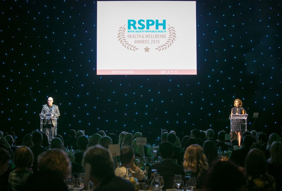 Zoomed out photo of two people on stage stood at lecterns at a previous RSPH Health & Wellbeing Awards with a large screen showing the RSPH logo.
