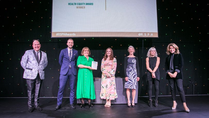 Seven people stood on stage with middle four posing with an Award with a background of a large screen with the RSPH logo in shot,