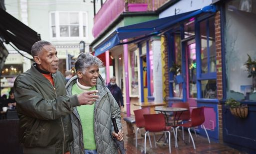 Older man and woman smiling while walking down a British high street on a rainy day with a traditional cafe in shot.