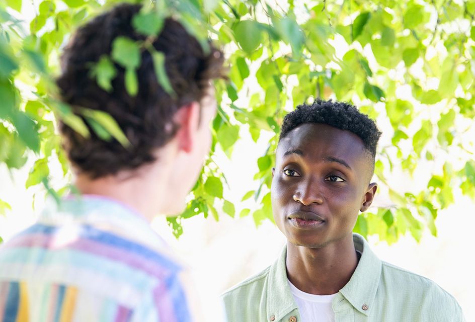 A man looking sympathetically at another man under a leafy tree on a sunny day.