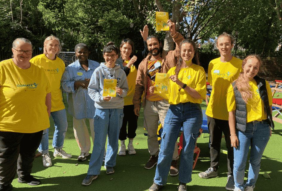 A group of eight North Paddington climate champions stands outdoors on a sunny day, smiling. The background features trees and colourful benches, conveying a cheerful, community-focused atmosphere.